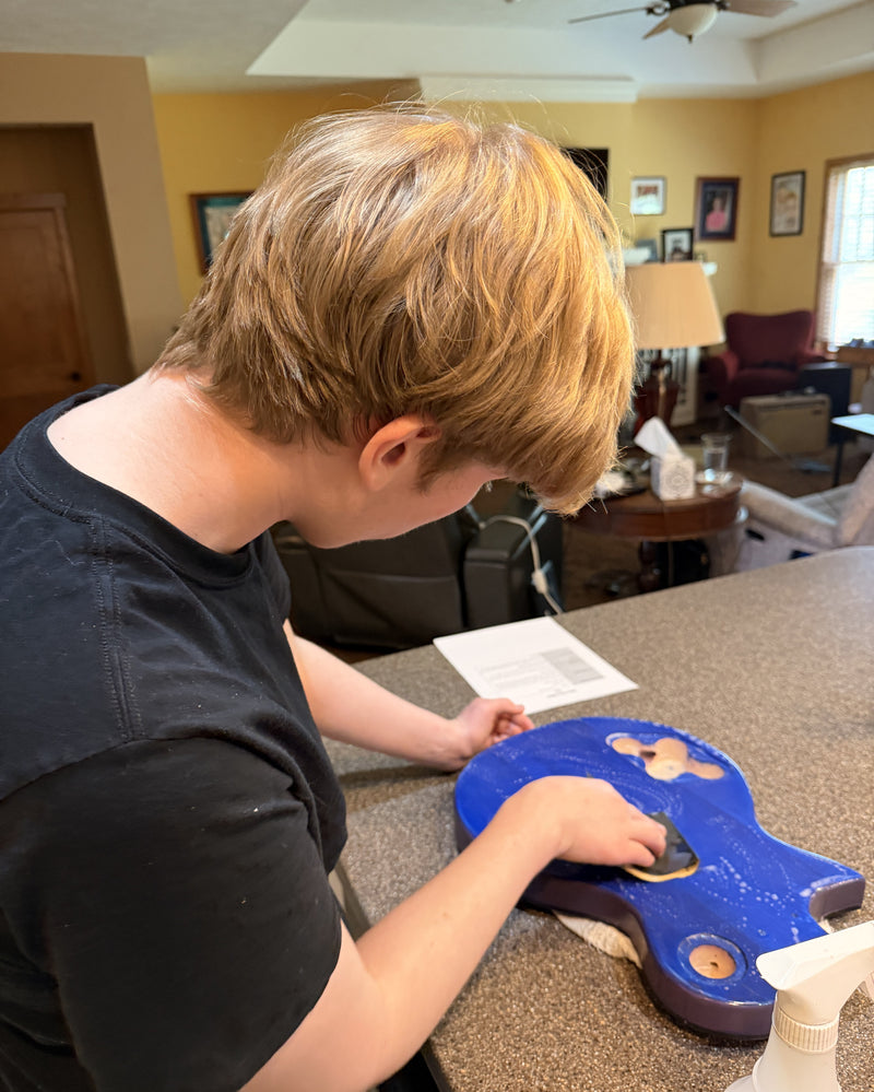 A first-time builder hand sanding the guitar body on a kitchen counter beside the kit instructions