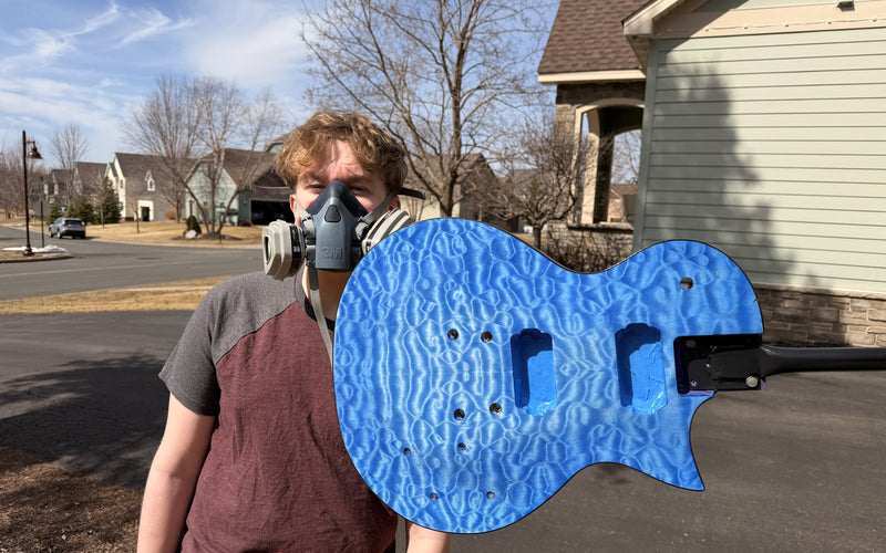Teenage builder in a respirator holding the quilted maple guitar body dyed bright blue, outdoors in the driveway before spraying