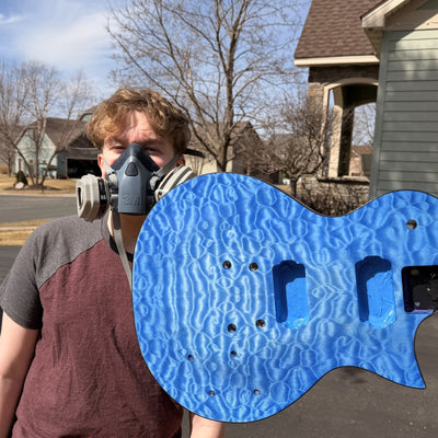 Teenage builder in a respirator holding the quilted maple guitar body dyed bright blue, outdoors in the driveway before spraying