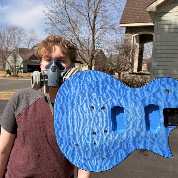Teenage builder in a respirator holding the quilted maple guitar body dyed bright blue, outdoors in the driveway before spraying