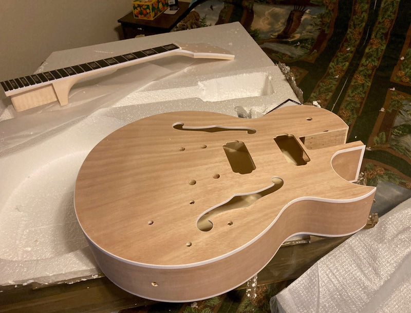 Unfinished semi-hollow guitar body with cream binding and f-holes, neck lying behind it in the box