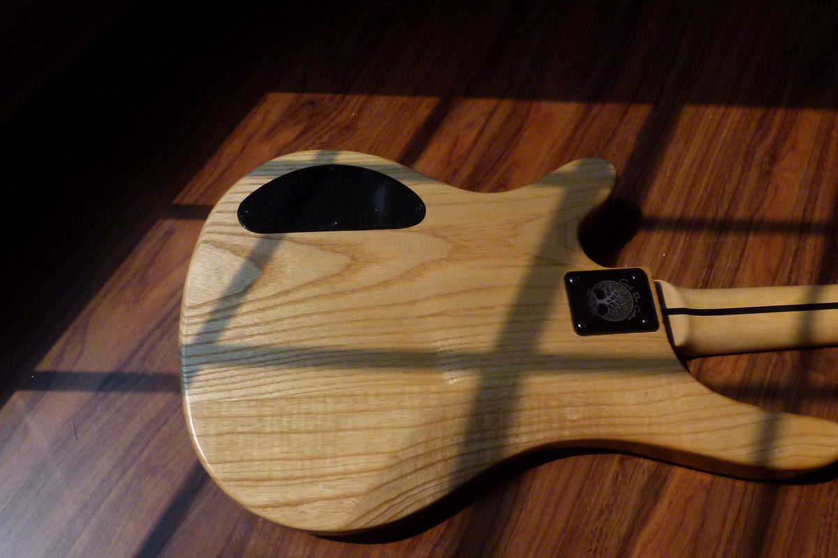 Back of the finished ash bass showing straight open grain, oval control cavity cover and an engraved black neck plate