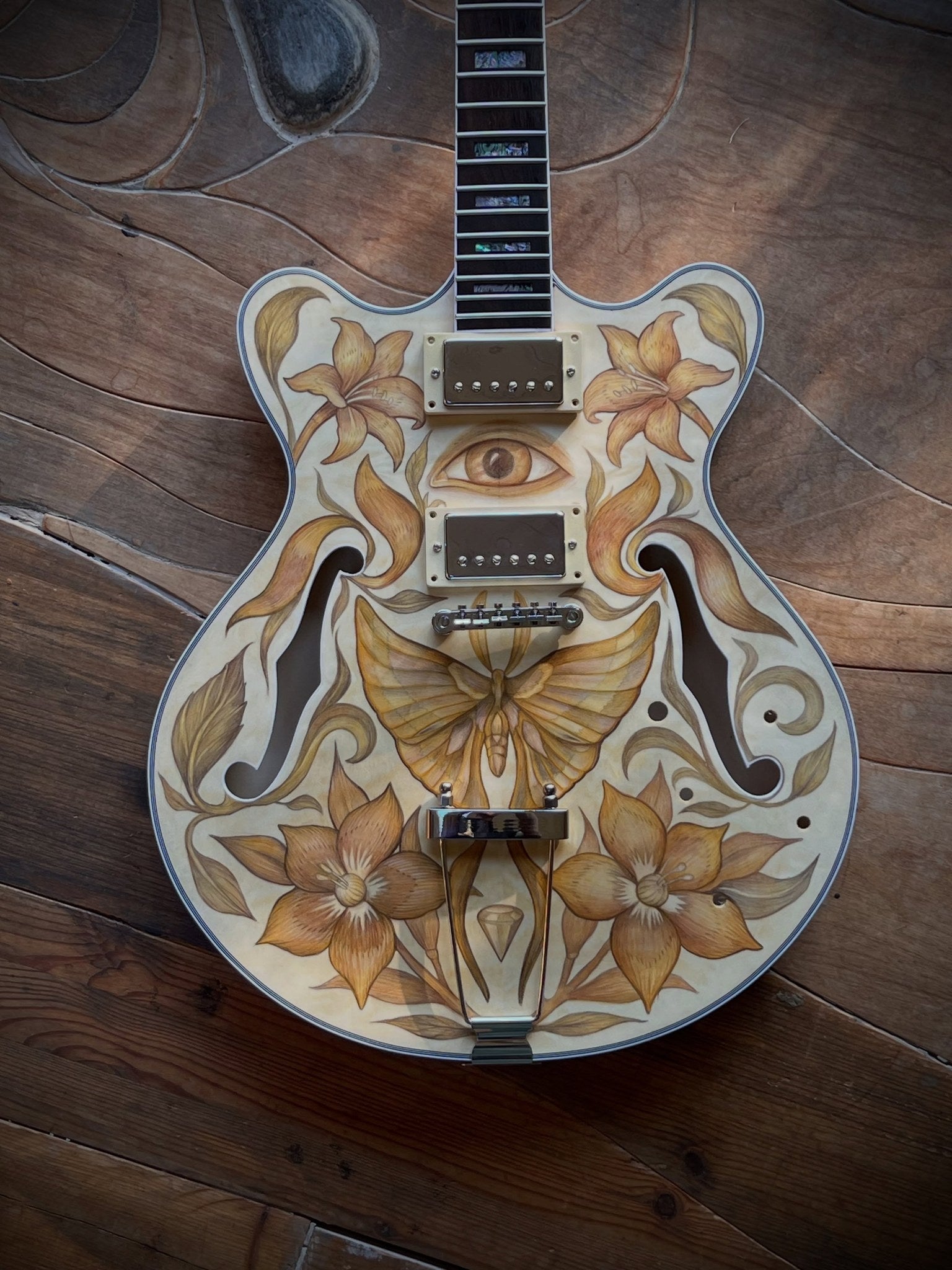 Full view of a semi-hollow double-cutaway guitar body with lilies, a moth and an eye drawn in colored pencil on bare flame maple, built by Peter-John de Villiers