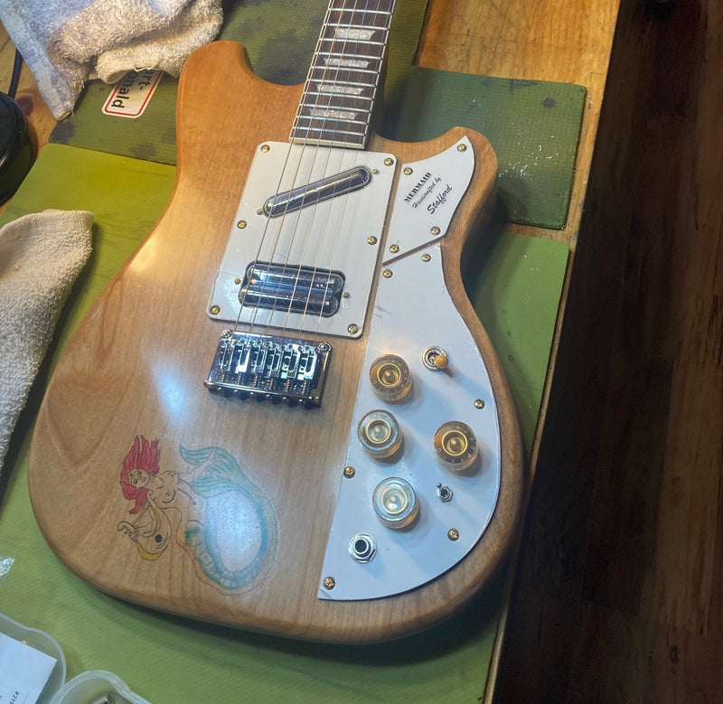 Natural wood guitar with a white pickguard, gold controls and a hand-drawn mermaid on the body