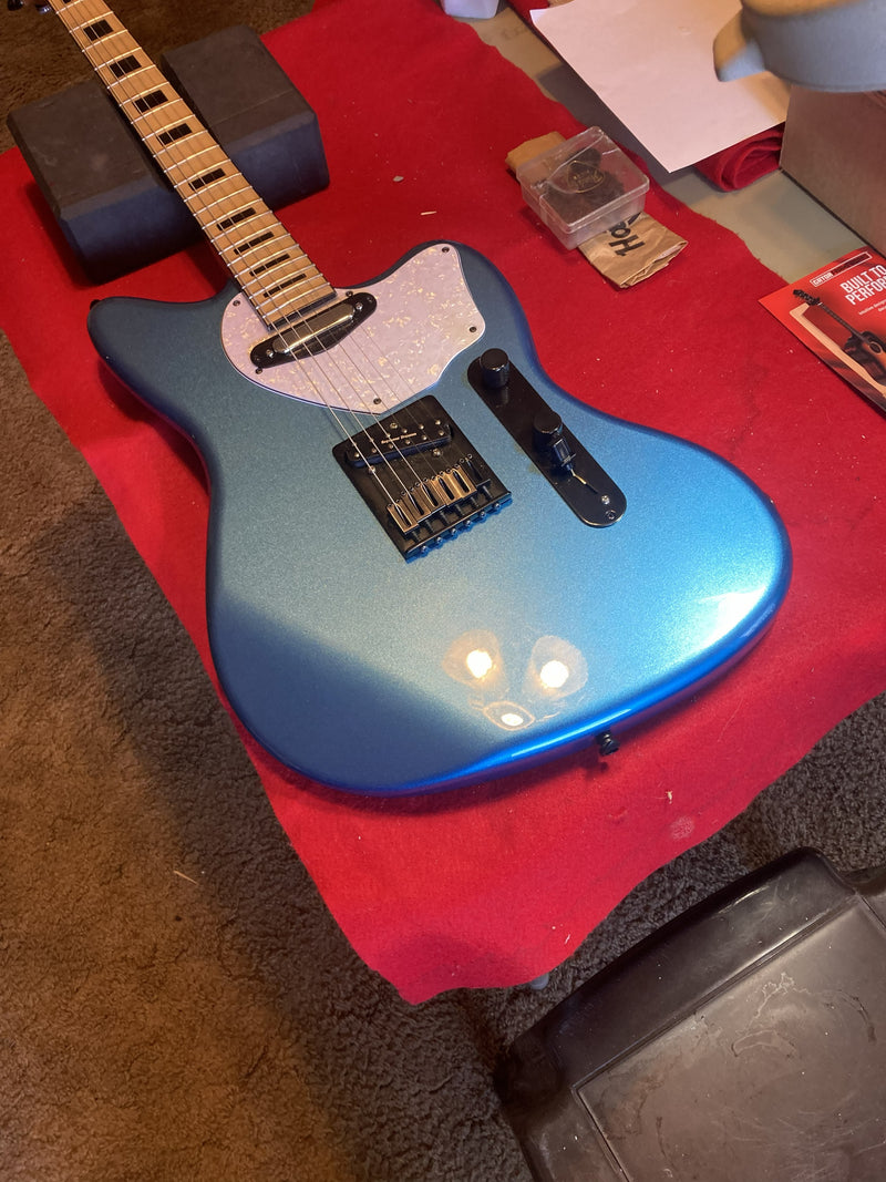 Metallic blue offset guitar with a maple neck and white pickguard, resting on a red cloth