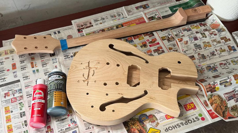 Raw guitar body with a rune freshly hand-carved into the top, stain cans on the newspaper beside it