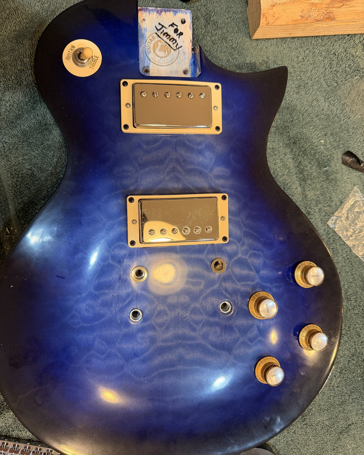 Assembled blue burst guitar body with gold humbucker rings and gold bell knobs, dedication written on the neck pocket block
