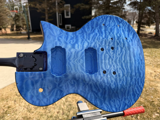 Blue dyed quilted maple guitar body clamped to a spraying stand outdoors