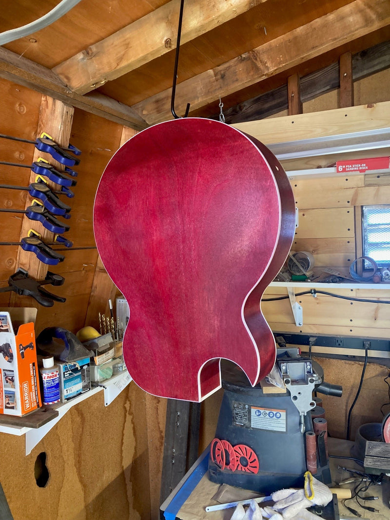 Red-stained semi-hollow guitar body hanging in a workshop after its sealer coat, clamps racked behind