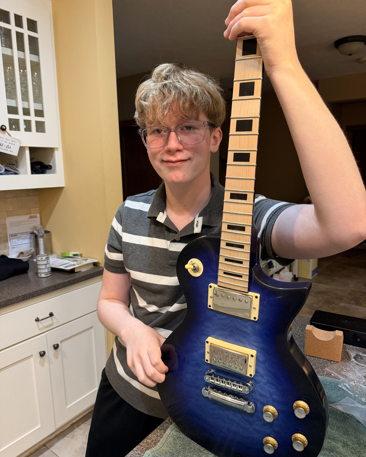 Teenager holding the completed blue burst L1 guitar with gold hardware and a maple block inlay fretboard