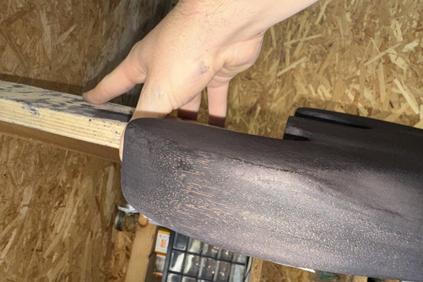 A builder holding the stained bass body up to daylight so the pale open pores read against the black